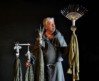 Man in monk like robe holds a rake, broom, and hoes that bear faces and clothes-like fabric